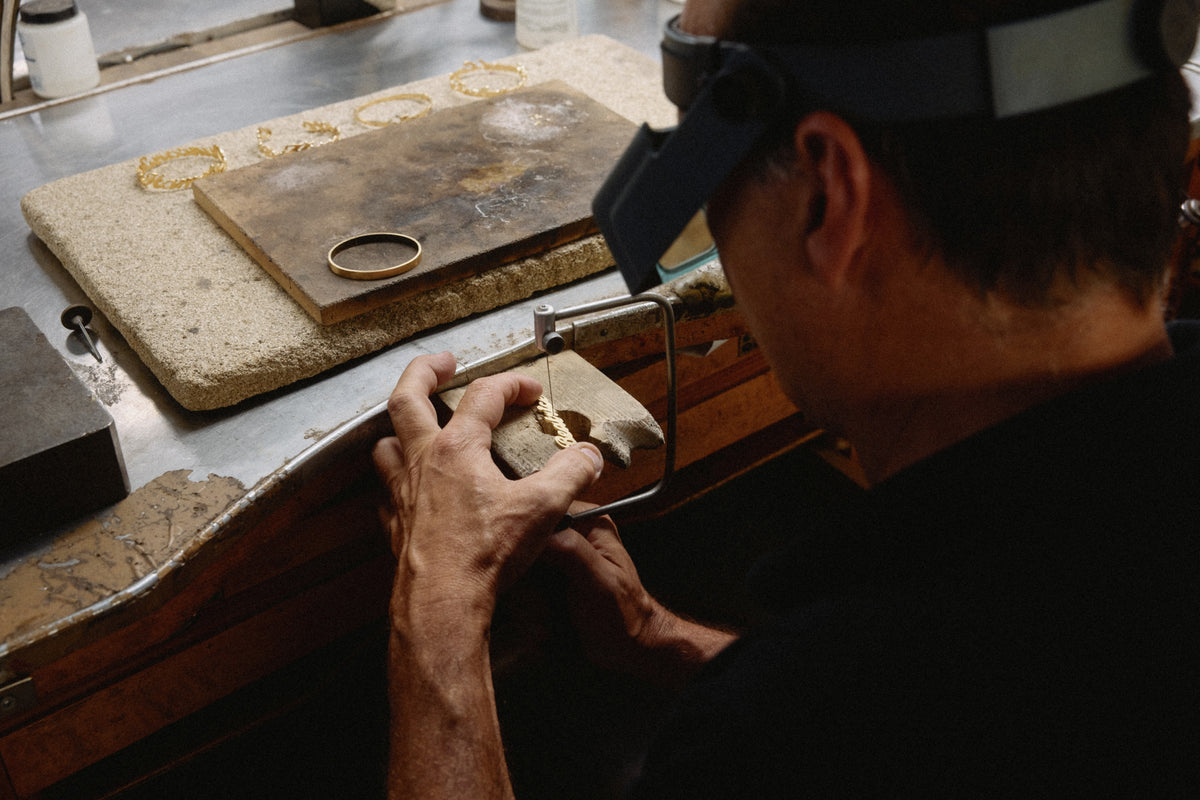 A goldsmith hand-saws gold letters at the workbench, with finished name bracelets laid out beside him
